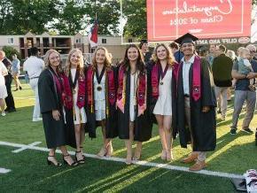 Six students in graduation regalia stand on the field under a sign reading "Congratulations Class of 2024"